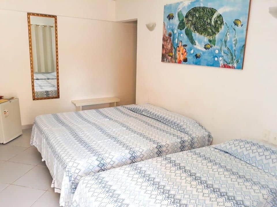Quarto da Pousada Santo Aleixo. Uma cama de casal e uma cama de solteiro do lado direito. No fundo, no canto esquerdo um frigobar, um espelho e um banco de madeira. Foto para ilustrar post sobre pousadas em Ipojuca.