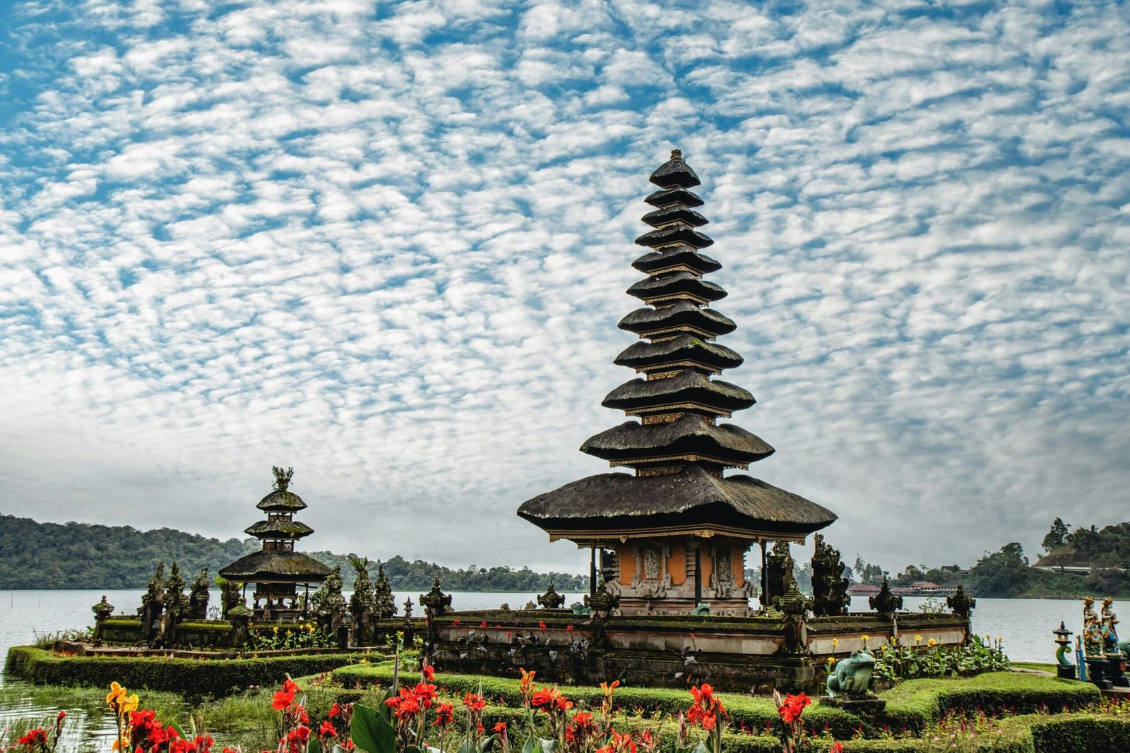 Templo Pura Ulun Danu Bratan com telhados de vários níveis é cercado por água e flores coloridas, sob um céu com nuvens dispersas. Representa Bali.