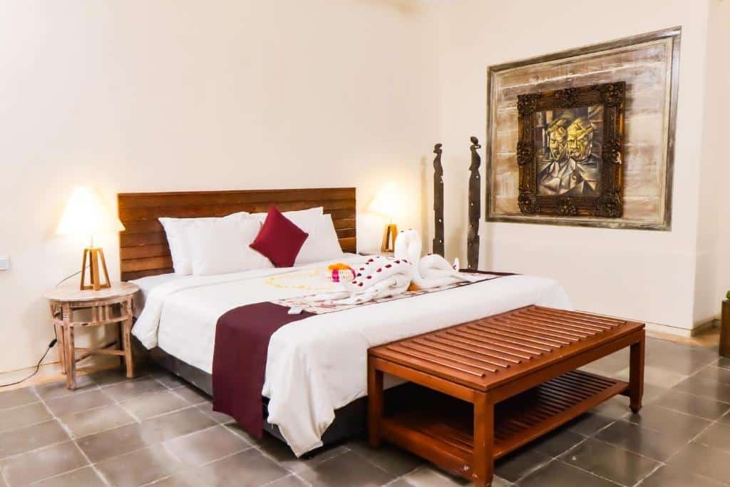 Quarto do Alam Puri Residence and Art Museum by BeeHive com cama de casal do lado esquerdo com uma cômoda em cada lado com luminária  e no pé da cama um banco de madeira. Representa Bali.