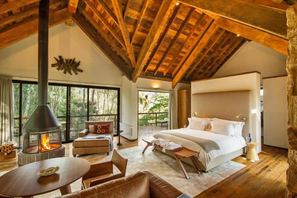 Quarto do Botanique Hotel & Spa com uma varanda ampla dando vista para árvores e montanhas, dentro o espaço é amplo e alto, há uma lareira, duas poltronas, chão de madeira, uma cama de casal e itens rústicos de decoração, para representar onde ficar em Campos do Jordão