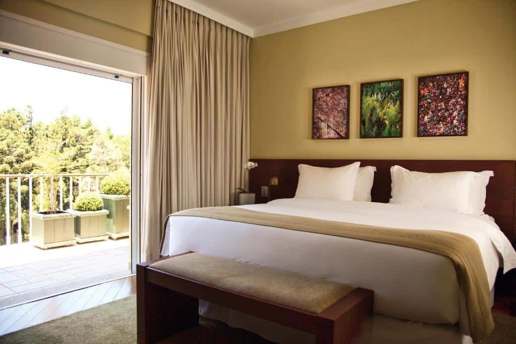 Quarto do Grande Hotel Campos do Jordao com uma sacada dando vista para o jardim e montanhas, dentro há uma cama de casal, e três quadros pequenos presos sob a cama