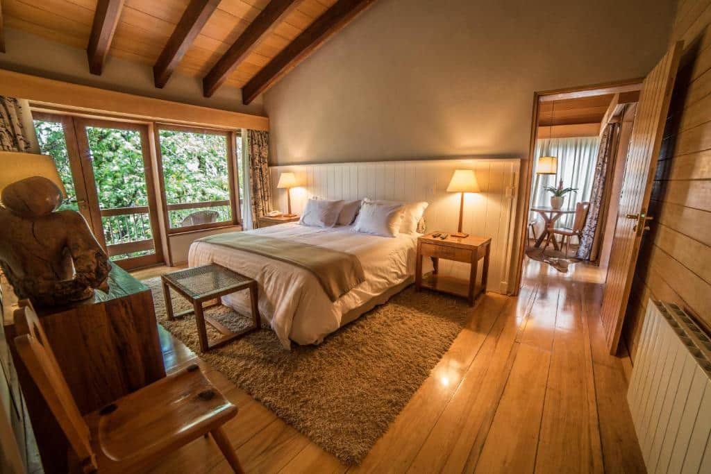 Quarto do Hotel Toriba com chão de madeira, sacada com vista para as montanhas, uma cama de casal, um tapete, duas cadeiras de madeira e duas mesinhas de cabeceira com abajures