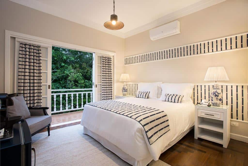 Quarto do Secreto Boutique Hotel com uma sacada com vista para as árvores, uma cama de casal, uma poltrona, um frigobar, chão de madeira com um tapete, há também duas mesinhas de cabeceira com abajures, para representar onde ficar em Campos do Jordão