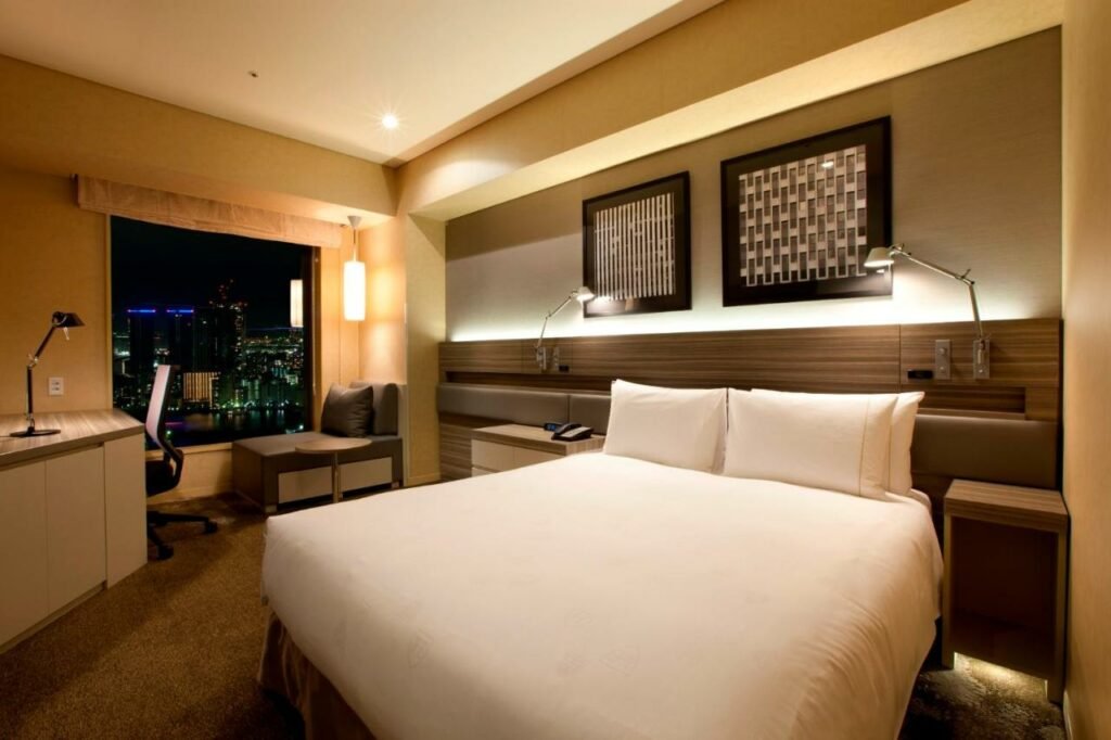 Quarto do hotel de Tóquio no Japão, The Royal Park Hotel Iconic Iconic Shiodome, com uma cama com edredom branco, carpete cinza, móveis em madeira, abajures e uma janela que tem a vista para a cidade. 