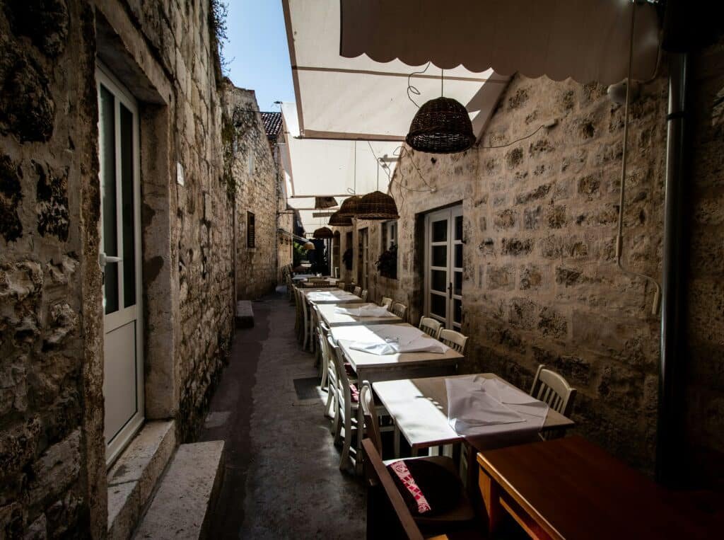 Beco estreito ao ar livre em Hvar, com paredes de pedra, ladeado por mesas e cadeiras brancas sob toldos, que leva a uma entrada distante. Representa o post de roteiro para Hvar.