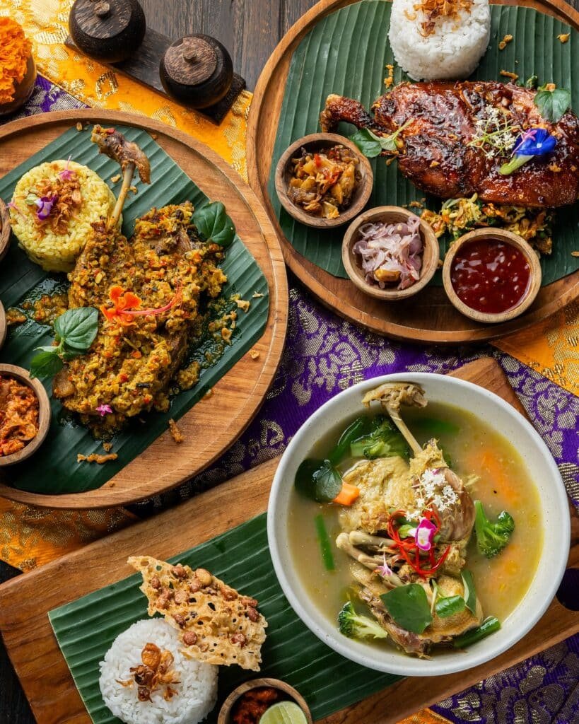 Uma variedade de pratos balineses em folhas de bananeira, incluindo frango grelhado, arroz, sopa de legumes e guarnições, em uma mesa de madeira. Ilustra a culinária balinesa no Sun Sun Warung.