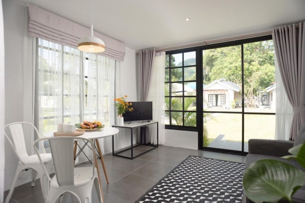 No Silan Residence, uma sala de estar aconchegante com uma mesa pequena, TV e uma janela grande. Uma porta de correr abre para um pátio com vista para casas e vegetação.