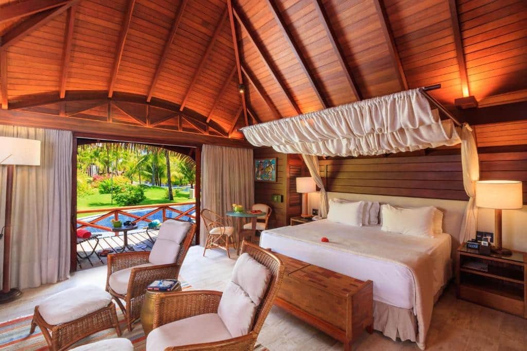 Quarto do NANNAI Muro Alto. Do lado direito uma mesa com duas cadeiras, uma cama de casal, com uma cômoda e um abajur de cada lado. Na frente um baú. Do lado esquerdo duas poltronas. No fundo, a varanda do quarto com vista para a piscina.