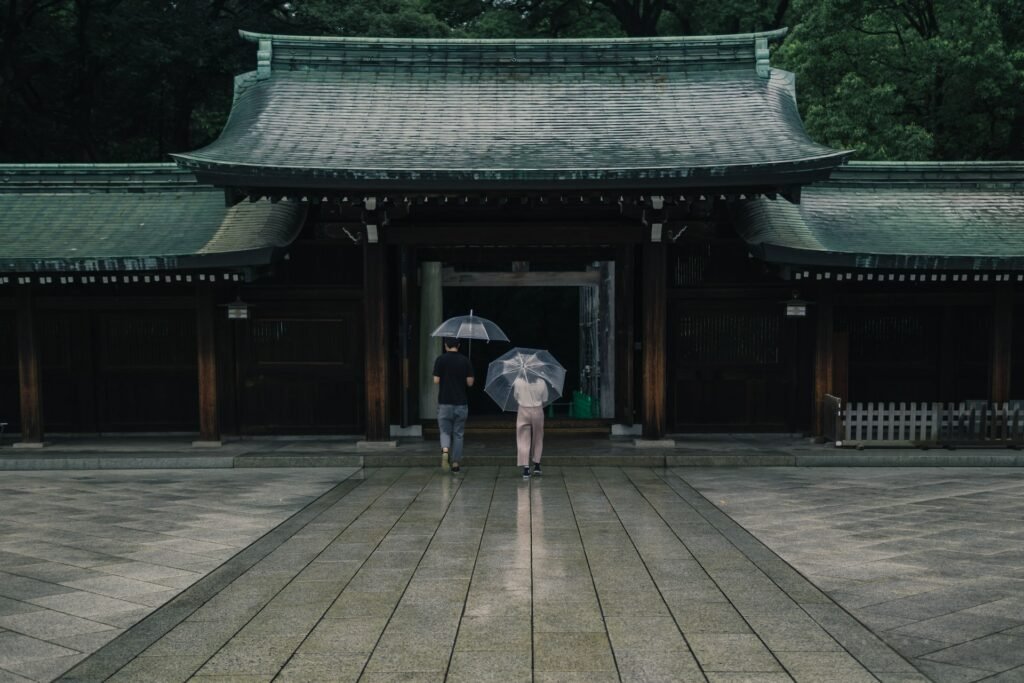 Duas pessoas com guarda-chuva entrando no templo de Menji Jingu, em dia de céu nublado e com chuva fina caindo, para representar o seguro viagem Tóquio. 