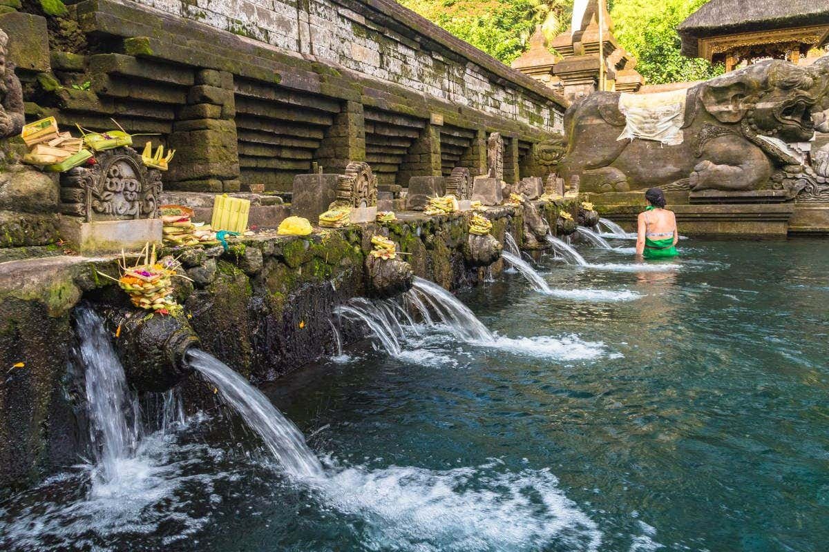 Uma pessoa em uma piscina com fontes de pedra, cercada por oferendas e esculturas, perto de um antigo muro de pedra. É o templo Tirta Empul.  Está ilustrando o post sobre o que fazer em Ubud.