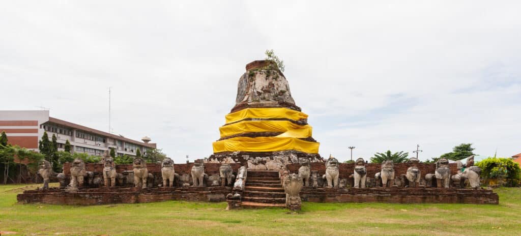 No Templo Thammikarat, antigo pagode de pedra envolto em tecido amarelo, cercado por estátuas de leões, com degraus de tijolos que levam até ele, em um campo gramado.
