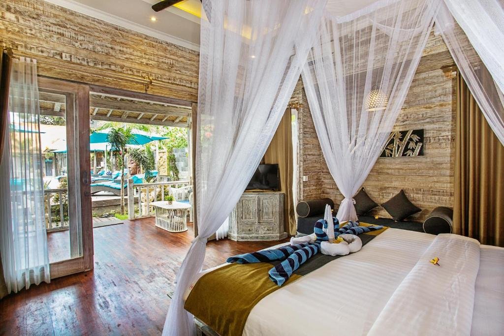 Um quarto rústico no hotel The Palm Groove Villas em Nusa Lembongan com cama de dossel, paredes de madeira e portas abertas que levam a um pátio com vista para a piscina e o jardim.