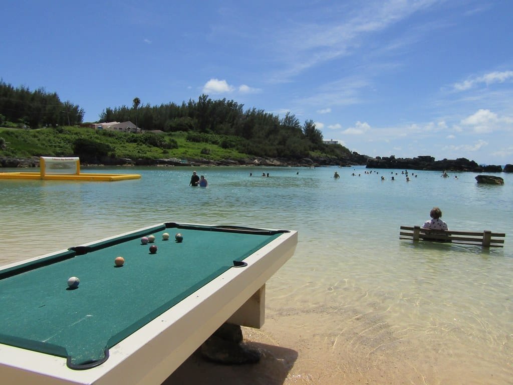 Uma mesa de sinuca no Tobacco Bay Beach perto de águas calmas, com pessoas nadando e sentadas em um banco na baía rasa. Imagem para ilustrar post sobre eSIM Bermudas.