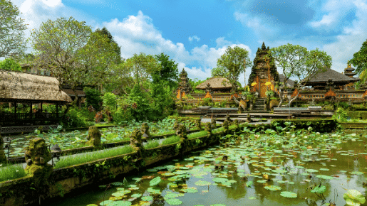Um lago cercado por plantas de lótus e arquitetura tradicional balinesa em Ubud, com árvores exuberantes e um céu azul ao fundo.