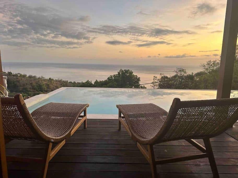 Imagem da estadia de Hillside Chalet with panoramic views com duas espreguiçadeiras ficam de frente para uma piscina de borda infinita com vista para o oceano ao pôr do sol, com árvores à direita e nuvens dispersas no céu.
