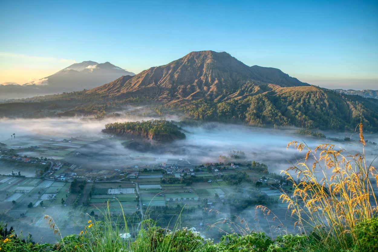 Uma vista panorâmica do Monte Batur com campos e vilas cobertos de neblina em primeiro plano, contra um céu azul claro.  Está ilustrando o post sobre o que fazer em Ubud.
