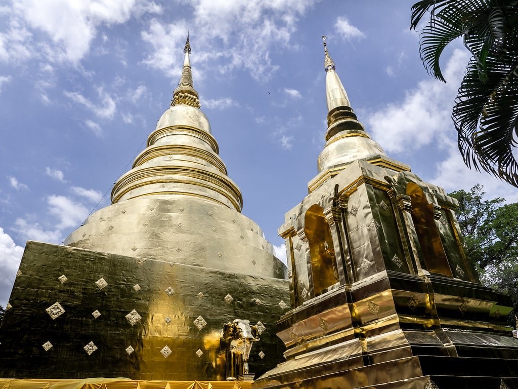 Pagodes dourados com desenhos intrincados e céu azul claro ao fundo, cercados por vegetação exuberante no 
Wat Phra Singh.